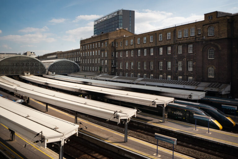 Paddington Station Platforms and the Historic GWR Train Shed 1 An elevated view of London Paddington Station shows the sweeping iron-and-glass train sheds designed in the mid-19th century for the Great Western Railway by Isambard Kingdom Brunel. The long canopies stretch over multiple platforms, where modern GWR Class 800 “Intercity Express” trains await departure. The adjacent red-brick offices of the original railway company still stand beside the terminus, a reminder of Paddington’s role as both a Victorian engineering triumph and a key link in Britain’s modern high-speed network.