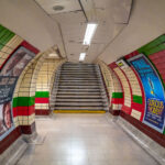 An underground corridor at London’s Piccadilly Circus Station connects the Bakerloo and Piccadilly line platforms deep beneath central London. Opened in 1906, the station sits directly under the city’s busiest intersection and handles tens of millions of passengers each year. The tunnels were rebuilt in the 1920s to improve crowd circulation, creating a complex network of escalators and passages like this one that channel travelers toward exits under Shaftesbury Avenue and Regent Street. Despite its age, the station remains one of the Underground’s most recognizable and heavily trafficked transport hubs in the West End.