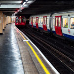 A pair of London Underground trains pass through Piccadilly Circus Station, one of the most recognizable stops in the network’s deep-level system. The curved platforms and distinctive cream-colored tilework reflect the 1920s modernization that gave the station its Art Deco influence, while the red, white, and blue train livery remains a symbol of the city’s transport identity. The platform’s polished floor and yellow safety line guide passengers along the busy interchange, located beneath the heart of London’s West End.