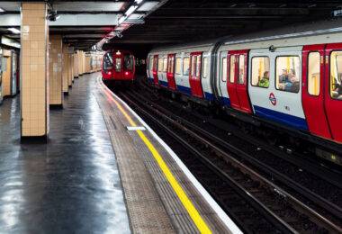 A pair of London Underground trains pass through Piccadilly Circus Station, one of the most recognizable stops in the network’s deep-level system. The curved platforms and distinctive cream-colored tilework reflect the 1920s modernization that gave the station its Art Deco influence, while the red, white, and blue train livery remains a symbol of the city’s transport identity. The platform’s polished floor and yellow safety line guide passengers along the busy interchange, located beneath the heart of London’s West End.