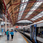 The platforms of London Victoria Station stretch beneath an ornate iron-and-glass roof, a hallmark of late 19th-century railway engineering. The intricate red and black trusses, designed for both strength and elegance, frame the soft natural light filtering through the skylights above. Commuters move toward the waiting trains of the Southern and Southeastern networks, a daily rhythm set against one of London’s most architecturally distinctive termini, where Victorian design continues to serve the modern rail era.