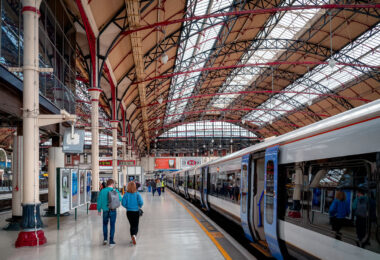 The platforms of London Victoria Station stretch beneath an ornate iron-and-glass roof, a hallmark of late 19th-century railway engineering. The intricate red and black trusses, designed for both strength and elegance, frame the soft natural light filtering through the skylights above. Commuters move toward the waiting trains of the Southern and Southeastern networks, a daily rhythm set against one of London’s most architecturally distinctive termini, where Victorian design continues to serve the modern rail era.
