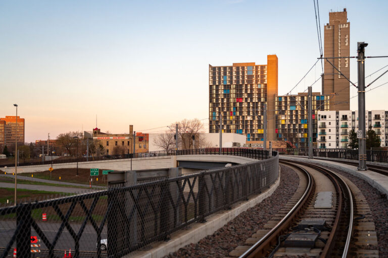 Light Rail Curve at Cedar–Riverside 4 The METRO Blue Line curves toward downtown Minneapolis near the Cedar–Riverside neighborhood, passing below the colorful concrete towers of Riverside Plaza at sunset. The complex, designed by architect Ralph Rapson and completed in 1973, is one of the city’s most distinctive examples of Brutalist modernism. This area, once part of the immigrant gateway to Minneapolis, now blends historic structures like the Mixed Blood Theatre with the transit corridor connecting neighborhoods across the city.