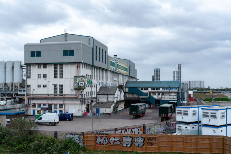 Tate & Lyle Silvertown Refinery, London Docklands 4 The Tate & Lyle sugar refinery in Silvertown remains one of the last major industrial landmarks on the north bank of the Thames. Established in 1878, the refinery was once the world’s largest sugar processing plant and continues operations today under the ASR Group, producing the famous Lyle’s Golden Syrup from its historic brick and steel complex.The building’s layered architecture—Edwardian masonry below, modern extensions above—reflects more than a century of adaptation to changing technologies and ownership. Despite the surrounding redevelopment of London’s Royal Docks into residential and commercial districts, this factory endures as a functioning reminder of the capital’s industrial heritage. The refinery’s green cat logo and “Out of the Strong Came Forth Sweetness” slogan have become enduring icons of British branding and trade history.
