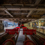 The buffer stops and terminating tracks beneath the iron spans of Paddington Station in London, photographed in the late afternoon light. Designed by Isambard Kingdom Brunel and opened in 1854, Paddington remains one of the city’s most architecturally significant rail termini. The exposed girders and riveted beams above reveal the original Victorian engineering still supporting modern operations, while the platforms now serve Elizabeth Line and Great Western Railway trains. The red hydraulic buffers and dense network of conduits illustrate the blend of 19th-century structure and 21st-century rail technology that defines Paddington’s evolution.