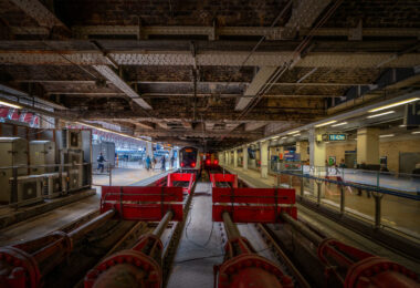 The buffer stops and terminating tracks beneath the iron spans of Paddington Station in London, photographed in the late afternoon light. Designed by Isambard Kingdom Brunel and opened in 1854, Paddington remains one of the city’s most architecturally significant rail termini. The exposed girders and riveted beams above reveal the original Victorian engineering still supporting modern operations, while the platforms now serve Elizabeth Line and Great Western Railway trains. The red hydraulic buffers and dense network of conduits illustrate the blend of 19th-century structure and 21st-century rail technology that defines Paddington’s evolution.