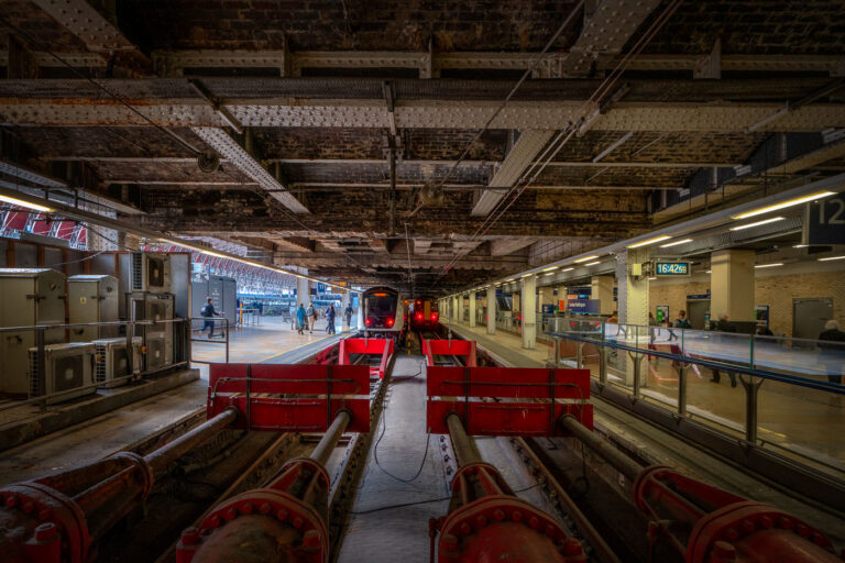 Terminus Tracks Beneath Paddington Station’s Ironwork 4 The buffer stops and terminating tracks beneath the iron spans of Paddington Station in London, photographed in the late afternoon light. Designed by Isambard Kingdom Brunel and opened in 1854, Paddington remains one of the city’s most architecturally significant rail termini. The exposed girders and riveted beams above reveal the original Victorian engineering still supporting modern operations, while the platforms now serve Elizabeth Line and Great Western Railway trains. The red hydraulic buffers and dense network of conduits illustrate the blend of 19th-century structure and 21st-century rail technology that defines Paddington’s evolution.