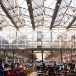 The Lawn at Paddington Station serves as the heart of the concourse, framed by the vast iron and glass canopy that defines Isambard Kingdom Brunel’s 19th-century design. Originally constructed in the 1850s, the station’s signature arched roof spans over the main platforms and the modernized public area below, now filled with cafés, travelers, and commuters. The space was reimagined in the early 2000s as part of a major restoration effort that preserved the Victorian ironwork while adding new steel and glass elements for natural light and openness. Beneath this intricate structure, passengers gather in a place where industrial heritage meets contemporary travel design.