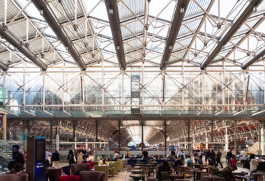 The Lawn at Paddington Station serves as the heart of the concourse, framed by the vast iron and glass canopy that defines Isambard Kingdom Brunel’s 19th-century design. Originally constructed in the 1850s, the station’s signature arched roof spans over the main platforms and the modernized public area below, now filled with cafés, travelers, and commuters. The space was reimagined in the early 2000s as part of a major restoration effort that preserved the Victorian ironwork while adding new steel and glass elements for natural light and openness. Beneath this intricate structure, passengers gather in a place where industrial heritage meets contemporary travel design.