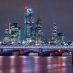 A night view across the River Thames captures Blackfriars Bridge illuminated in cool tones beneath the glass towers of London’s financial district. The skyline features some of the city’s most recognizable modern landmarks — including the Leadenhall Building (the “Cheesegrater”), 22 Bishopsgate, and the distinctive curved façade of 20 Fenchurch Street, known as the “Walkie-Talkie.” Red aviation lights and construction cranes trace the ongoing vertical growth of the Square Mile, while reflections shimmer on the Thames below. The composition underscores the contrast between Victorian bridge engineering and the contemporary architecture reshaping London’s historic core.