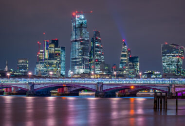 A night view across the River Thames captures Blackfriars Bridge illuminated in cool tones beneath the glass towers of London’s financial district. The skyline features some of the city’s most recognizable modern landmarks — including the Leadenhall Building (the “Cheesegrater”), 22 Bishopsgate, and the distinctive curved façade of 20 Fenchurch Street, known as the “Walkie-Talkie.” Red aviation lights and construction cranes trace the ongoing vertical growth of the Square Mile, while reflections shimmer on the Thames below. The composition underscores the contrast between Victorian bridge engineering and the contemporary architecture reshaping London’s historic core.