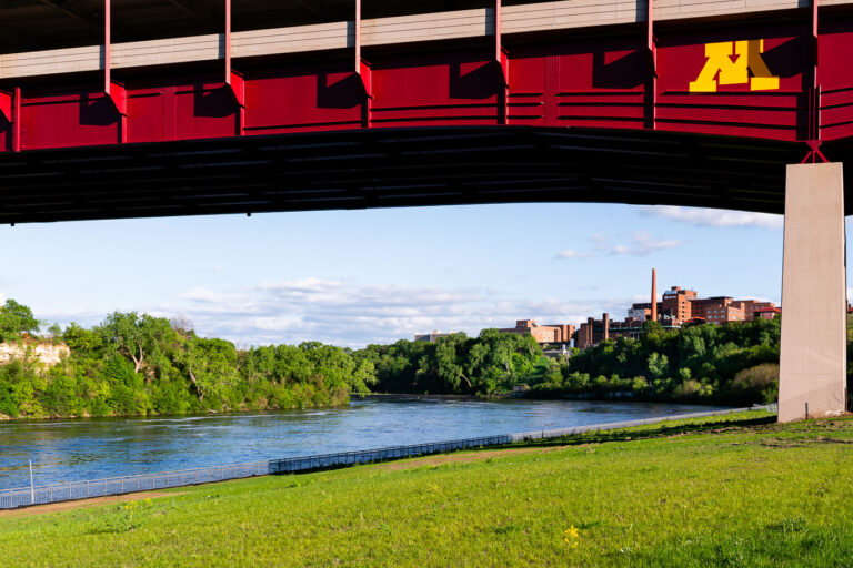 Mississippi River Beneath the Washington Avenue Bridge 1 The maroon and gold Washington Avenue Bridge spans the Mississippi River, linking the University of Minnesota’s East and West Bank campuses. Seen from below, the bridge’s bold steel trusses frame the calm river and limestone bluffs, while the university’s West Bank buildings rise in the background. The bridge, completed in 1965, carries both vehicular and pedestrian traffic—including the campus’s light rail line—and serves as one of the most recognizable landmarks connecting the university’s two halves across the river.