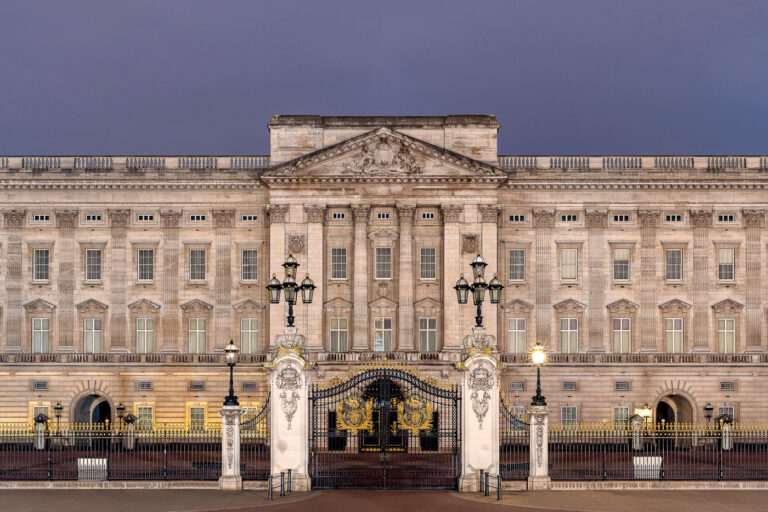 Buckingham Palace Front Facade at Dusk 2 The east front of Buckingham Palace, photographed at dusk, displays the neoclassical grandeur of Britain’s royal residence. Designed by architect Sir Aston Webb and completed in 1913, the symmetrical limestone facade is illuminated softly against a violet evening sky. Ornate wrought-iron gates adorned with gilded royal crests frame the main entrance, while gas-style lamps evoke the palace’s Edwardian-era detailing. The balanced rhythm of Corinthian pilasters and sash windows underscores the building’s enduring place as both a functioning royal residence and an architectural symbol of the British monarchy.