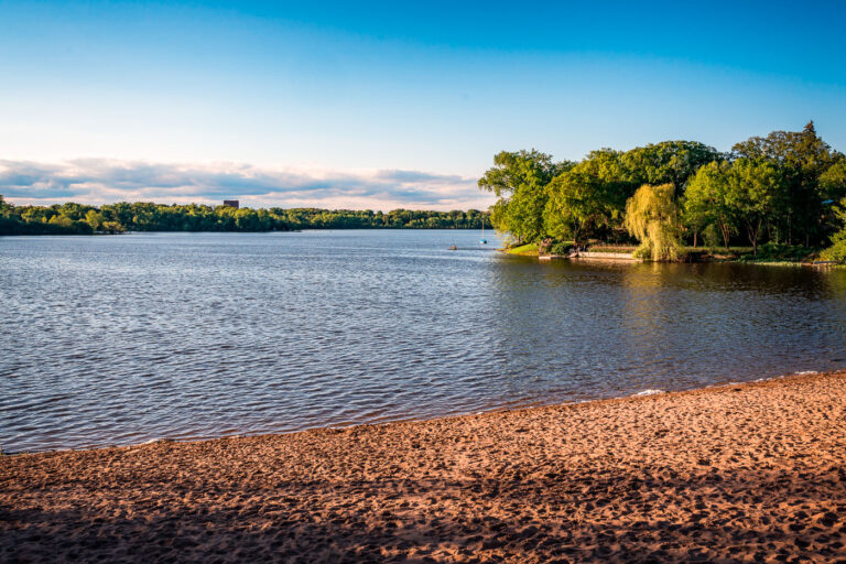 Cedar Lake South Beach in Evening Light 1 Cedar Lake South Beach in Minneapolis, photographed in the evening light, captures one of the city’s quieter public beaches along the Chain of Lakes. Located west of Bde Maka Ska, Cedar Lake is known for its natural shoreline and tree-lined surroundings, offering a more secluded experience compared to the busier neighboring lakes. The sandy beach, calm water, and sailboats anchored offshore highlight Minneapolis’s integration of recreation, nature, and urban life within a few miles of downtown.