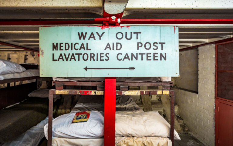 Clapham North Deep Shelter Sign: Medical Aid, Lavatories, Canteen 1 A directional sign for "Medical Aid Post, Lavatories, Canteen" points the way within the Clapham North Deep Shelter. Constructed between 1940 and 1942, this subterranean bunker was one of eight built beneath London's Underground to protect civilians during World War II air raids. The sign highlights the essential services provided in these deep-level shelters, which offered a self-contained environment for thousands of Londoners. The presence of bunk beds, as seen in the foreground, underscores the shelter's role as a temporary home during the Blitz, serving as a stark reminder of wartime resilience.