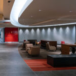 Collaborative Lounge at Target Corporate Headquarters 3 An interior lounge and corridor at Target Corporation’s headquarters in Minneapolis, designed to encourage informal meetings, employee collaboration, and visitor reception. The space reflects Target’s post-2010 modern workplace philosophy — blending minimalist corporate architecture with hospitality-inspired interiors. Curved ceiling soffits, accent lighting, and red brand-themed wall panels soften the otherwise sleek office environment, while modular seating and large communal tables support group discussions and mobile work. This type of open, flexible workspace became increasingly common in large corporate campuses in the 2010s, as U.S. companies shifted away from traditional cubicle layouts toward collaborative, design-driven environments influenced by retail and tech sectors. The Target brand’s signature red is subtly integrated throughout, reinforcing corporate identity within the workspace’s architecture.