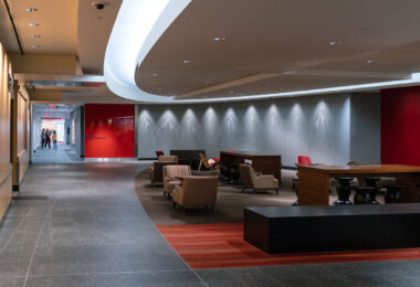 An interior lounge and corridor at Target Corporation’s headquarters in Minneapolis, designed to encourage informal meetings, employee collaboration, and visitor reception. The space reflects Target’s post-2010 modern workplace philosophy — blending minimalist corporate architecture with hospitality-inspired interiors. Curved ceiling soffits, accent lighting, and red brand-themed wall panels soften the otherwise sleek office environment, while modular seating and large communal tables support group discussions and mobile work. This type of open, flexible workspace became increasingly common in large corporate campuses in the 2010s, as U.S. companies shifted away from traditional cubicle layouts toward collaborative, design-driven environments influenced by retail and tech sectors. The Target brand’s signature red is subtly integrated throughout, reinforcing corporate identity within the workspace’s architecture.