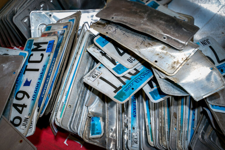 Recycled License Plates at K&K Metals 3 Stacks of discarded Minnesota license plates sit ready for recycling at K&K Metals in Minneapolis. The familiar “10,000 Lakes” slogan and blue gradient design peek through layers of grime and bent aluminum, remnants of years of use on the road. Facilities like this one reclaim the aluminum from expired or damaged plates, melting it down for reuse—part of a statewide effort to reduce waste and recover valuable materials from the transportation system.