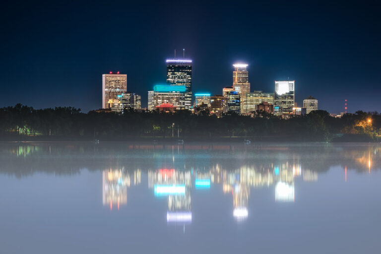 Fog Over Bde Maka Ska with the Minneapolis Skyline 2 The illuminated Minneapolis skyline rises behind a fog-covered Bde Maka Ska, its towers mirrored in the lake’s still surface. Buildings like the IDS Center, Capella Tower, and Wells Fargo Center shine through the mist, their neon accents reflecting off the calm water. Captured at night, the photograph contrasts the cool, modern glow of downtown with the serene, natural quiet of the city’s largest lake—a reminder of how Minneapolis balances its urban form with its deep connection to water and landscape.