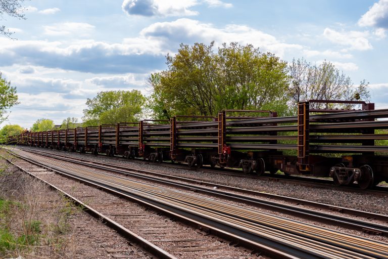 Rail Cars Hauling New Track for the Green Line Extension 4 Long train of empty freight cars lined up on railroad tracks beside green spring vegetation.