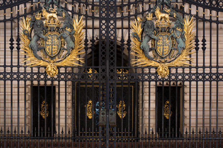 Buckingham Palace Gates and Royal Coat of Arms 4 The ornate front gates of Buckingham Palace display the Royal Coat of Arms of the United Kingdom, rendered in gilded bronze against black wrought iron. The emblem—featuring a crowned lion and a chained unicorn supporting a quartered shield—symbolizes the unity of England, Scotland, and Ireland under the Crown. Each detail of the design, from the gilded laurel bursts to the scrollwork on the surrounding ironwork, reflects both heraldic tradition and the craftsmanship of early twentieth-century metalworkers. These gates form one of the most photographed entrances in the world, standing as a ceremonial threshold to Britain’s royal residence.