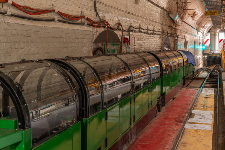 London Post Office Railway – Mail Rail at the Postal Museum 1 Beneath the streets of central London lies one of the city’s lesser-known engineering feats—the Post Office Railway, more commonly known as the Mail Rail. Built in the early 20th century and officially opened in 1927, the driverless electric railway carried letters and parcels between key sorting offices and railway stations, including Paddington, Mount Pleasant, and Liverpool Street. The tunnels, only 2 feet wide and running for over six miles, allowed mail to move across the capital in a fraction of the time it took above ground, unaffected by London’s constant congestion.
The trains, such as the preserved unit pictured here, were designed to operate automatically, hauling specially built mail containers along narrow tracks deep below the city. Each train could carry up to 12 tons of post at speeds of up to 40 miles per hour, operating 22 hours a day at the network’s peak. The system remained in service until 2003, when changing logistics and new road systems made it redundant.
Today, visitors to the Postal Museum in Clerkenwell can ride a restored section of the railway and see the machinery that once kept London’s communication network in motion. The preserved infrastructure—brick tunnels, signal systems, and compact rolling stock—stands as a rare example of early automation in transport history, reflecting Britain’s long-standing innovation in engineering and urban logistics.