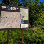 A detour sign for the Southwest Light Rail Transit (SWLRT) project stands beside a bike trail in Minneapolis, showing the rerouted path for cyclists and pedestrians during construction of the Green Line Extension. The detailed map outlines temporary closures through the Kenilworth Corridor and Cedar Lake areas, where new light rail tracks are being installed to connect downtown Minneapolis with the southwestern suburbs. The project, one of the largest public works efforts in Minnesota history, has significantly altered popular urban trails like the Midtown Greenway and Cedar Lake Trail since construction began in the late 2010s.