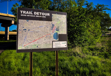A detour sign for the Southwest Light Rail Transit (SWLRT) project stands beside a bike trail in Minneapolis, showing the rerouted path for cyclists and pedestrians during construction of the Green Line Extension. The detailed map outlines temporary closures through the Kenilworth Corridor and Cedar Lake areas, where new light rail tracks are being installed to connect downtown Minneapolis with the southwestern suburbs. The project, one of the largest public works efforts in Minnesota history, has significantly altered popular urban trails like the Midtown Greenway and Cedar Lake Trail since construction began in the late 2010s.