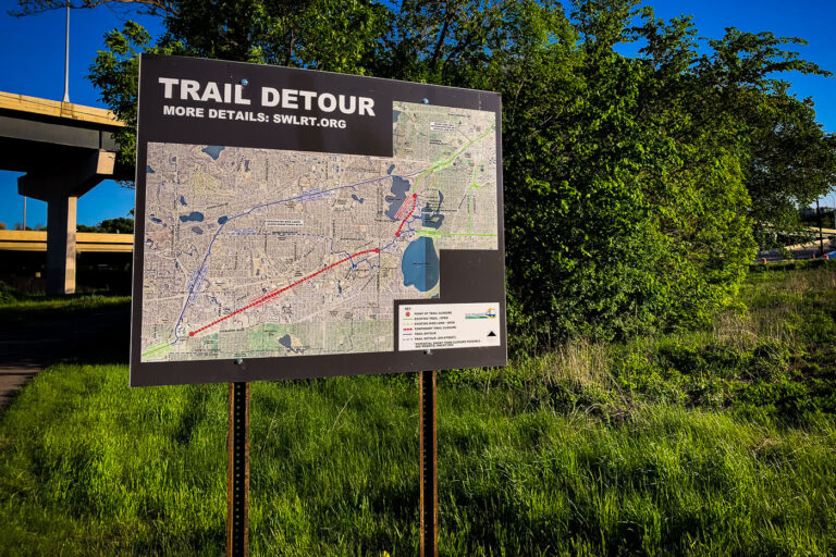 Southwest LRT Trail Detour Sign, Minneapolis 1 A detour sign for the Southwest Light Rail Transit (SWLRT) project stands beside a bike trail in Minneapolis, showing the rerouted path for cyclists and pedestrians during construction of the Green Line Extension. The detailed map outlines temporary closures through the Kenilworth Corridor and Cedar Lake areas, where new light rail tracks are being installed to connect downtown Minneapolis with the southwestern suburbs. The project, one of the largest public works efforts in Minnesota history, has significantly altered popular urban trails like the Midtown Greenway and Cedar Lake Trail since construction began in the late 2010s.