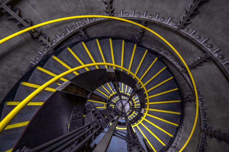 Spiral Emergency Staircase at Clapham North Underground Station 3 A view inside one of the distinctive spiral staircases of Clapham North Underground Station on the Northern Line, part of London’s deep-level tube network completed in the 1920s. The cast-iron stairwell descends nearly 120 feet below ground, wrapping tightly around a central ventilation column used to regulate air pressure and temperature within the tunnels. The staircase, equipped with yellow anti-slip edges and a steel handrail, was designed as an emergency exit and maintenance access route for the deep-bore platforms below.Stations like Clapham North, along with its twin Clapham Common, are known for their rare narrow island platforms and compact circular shafts — relics of early tube engineering when space and construction methods were limited. The robust riveted metalwork and industrial geometry of the stairwell reflect the period’s emphasis on function and endurance, forming part of London’s complex subterranean infrastructure still in use more than a century later.