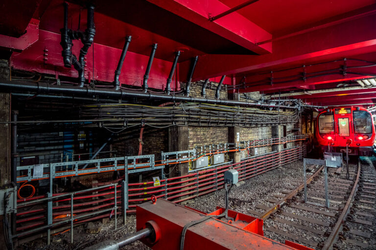 Baker Street Station – Subsurface Track and Signal Infrastruct 3 Deep beneath central London, Baker Street’s Metropolitan Line platforms reveal the layered engineering of one of the oldest functioning railway systems in the world. The exposed brick tunnel and steel framing date back to the Victorian “cut-and-cover” era of the 1860s, when steam locomotives first ran through these very corridors. The heavy red girders seen above were reinforced during modern refurbishments, supporting the city streets above while housing utilities and cable conduits that power today’s Underground network.
The train at the far end belongs to the S8 Stock series, introduced in 2010 by Bombardier for the Metropolitan Line, equipped with air conditioning and regenerative braking. The dense web of cables and control boxes along the wall carries signal, communication, and traction power circuits, all vital to the line’s safe operation. Baker Street’s infrastructure embodies London Transport’s continual evolution—from soot-covered tunnels to precision-controlled, electrically powered systems still running along the same 19th-century alignments.