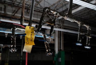 Inside an abandoned industrial building, a set of corroded water valves and pipes hangs overhead, marked with a fading yellow tag reading “City Water.” The scene captures the quiet afterlife of urban infrastructure—where once-busy mechanical systems now sit dormant beneath exposed rafters and dim fluorescent tubes. These remaining fixtures, wrapped in aging insulation and patched tape, reveal the skeletal logic of a space once dependent on pressurized networks, maintenance routines, and municipal supply lines that connected the factory to the city’s hidden circulatory systems.
