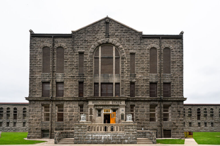 Wisconsin State Reformatory Main Entrance – Green Bay, Wiscons 2 Built in 1898, the Wisconsin State Reformatory in Green Bay is a monumental example of early 20th-century correctional architecture, designed to project authority through its Romanesque Revival stonework and fortress-like symmetry. The façade, constructed from locally quarried gray limestone, features narrow arched windows, heavy masonry, and a commanding central pediment above the main entryway. Originally intended as a progressive institution focused on rehabilitation rather than punishment, the reformatory combined rigid design with reform-era ideals. The weathered sign above the entrance—still reading “Wisconsin State Reformatory”—marks one of the few remaining intact examples of this architectural period in the state’s penal history.