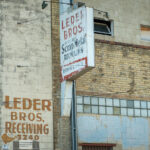 The weathered brick façade of Leder Bros. Scrap Metal Recycling reflects a century of small-scale industrial life in north Minneapolis. Faded hand-painted lettering and a rusting mid-century sign mark the receiving entrance for what was once a busy recycling yard, a place where scrap from the city’s rail lines and factories found new purpose. The combination of concrete block, yellow brick, and glass block windows speaks to a utilitarian design typical of the post-war industrial boom. Today, its aging surfaces and surveillance cameras evoke a district in transition, where traces of heavy industry linger amid redevelopment and adaptive reuse.