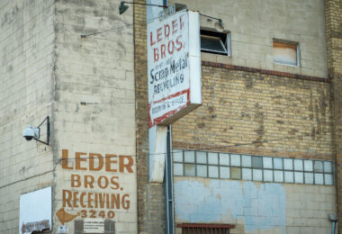 The weathered brick façade of Leder Bros. Scrap Metal Recycling reflects a century of small-scale industrial life in north Minneapolis. Faded hand-painted lettering and a rusting mid-century sign mark the receiving entrance for what was once a busy recycling yard, a place where scrap from the city’s rail lines and factories found new purpose. The combination of concrete block, yellow brick, and glass block windows speaks to a utilitarian design typical of the post-war industrial boom. Today, its aging surfaces and surveillance cameras evoke a district in transition, where traces of heavy industry linger amid redevelopment and adaptive reuse.