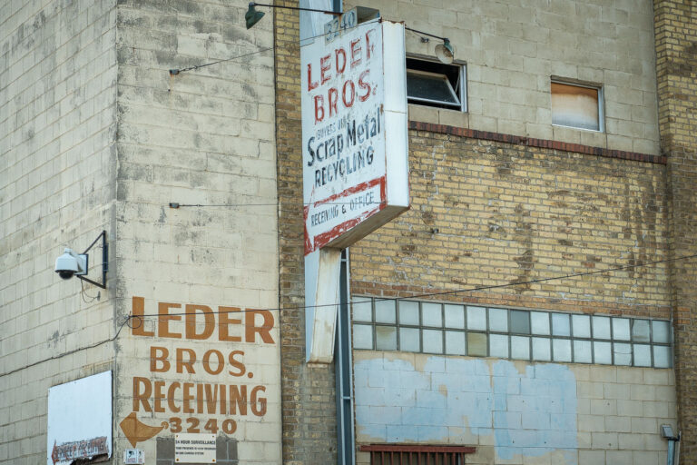 Leder Bros. Scrap Metal – North Minneapolis Industrial Heritag 2 The weathered brick façade of Leder Bros. Scrap Metal Recycling reflects a century of small-scale industrial life in north Minneapolis. Faded hand-painted lettering and a rusting mid-century sign mark the receiving entrance for what was once a busy recycling yard, a place where scrap from the city’s rail lines and factories found new purpose. The combination of concrete block, yellow brick, and glass block windows speaks to a utilitarian design typical of the post-war industrial boom. Today, its aging surfaces and surveillance cameras evoke a district in transition, where traces of heavy industry linger amid redevelopment and adaptive reuse.