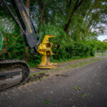 A tracked John Deere feller buncher with a yellow cutting head sits idle beside the Cedar Lake Trail in St. Louis Park, Minnesota, during early site preparation for the Southwest Light Rail Transit (LRT) extension. The machine’s “KEEP BACK 300 FT / 90 M” arm warning and the trail’s closed fencing mark the transition from a popular cycling route to an active construction corridor. In the distance, the bridge approach and industrial structures hint at the scale of redevelopment that reshaped the rail and trail landscape through Minneapolis’s western suburbs.