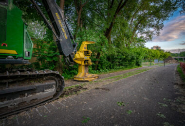 A tracked John Deere feller buncher with a yellow cutting head sits idle beside the Cedar Lake Trail in St. Louis Park, Minnesota, during early site preparation for the Southwest Light Rail Transit (LRT) extension. The machine’s “KEEP BACK 300 FT / 90 M” arm warning and the trail’s closed fencing mark the transition from a popular cycling route to an active construction corridor. In the distance, the bridge approach and industrial structures hint at the scale of redevelopment that reshaped the rail and trail landscape through Minneapolis’s western suburbs.