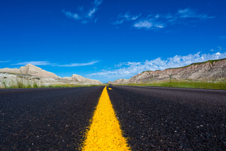Highway 44 Through the Badlands 2 A newly paved stretch of Highway 44 cuts through the rugged formations near Scenic, South Dakota, beneath a vivid summer sky. This east–west route skirts the southern boundary of Badlands National Park, where blacktop meets eroded buttes carved by millennia of wind and water. The low-angle view emphasizes the contrast between the engineered precision of the highway and the raw geologic textures of the surrounding landscape—an enduring symbol of travel through one of the Great Plains’ most striking natural corridors.
