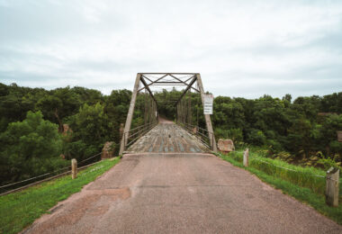 An old steel truss bridge spans Split Rock Creek near the town of Garretson, South Dakota, carrying a narrow, weathered wooden deck over the rocky gorge below. Built in the early 20th century, this Pratt through truss design reflects an era when such riveted steel frameworks connected small Midwestern towns before the advent of modern concrete spans. Its utilitarian geometry and riveted latticework remain intact, framed by the lush, tree-lined valley of Split Rock Creek—a setting that once hosted flour mills, quarries, and river crossings essential to the region’s early development. The bridge now stands as a quiet relic of rural American engineering, linking history and landscape on the edge of the prairie.
