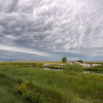 An approaching storm looms over the rolling grasslands near Interior, South Dakota, at the edge of Badlands National Park. The layered clouds form dramatic bands above the wetland and open prairie, where a small structure and fence line mark the human presence in this otherwise vast and untamed landscape. From this vantage point near the White River, the eroded formations of the Badlands rise faintly in the distance, emphasizing the contrast between the lush foreground and the arid, sculpted terrain beyond.