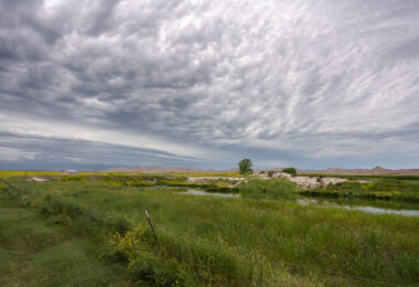 An approaching storm looms over the rolling grasslands near Interior, South Dakota, at the edge of Badlands National Park. The layered clouds form dramatic bands above the wetland and open prairie, where a small structure and fence line mark the human presence in this otherwise vast and untamed landscape. From this vantage point near the White River, the eroded formations of the Badlands rise faintly in the distance, emphasizing the contrast between the lush foreground and the arid, sculpted terrain beyond.