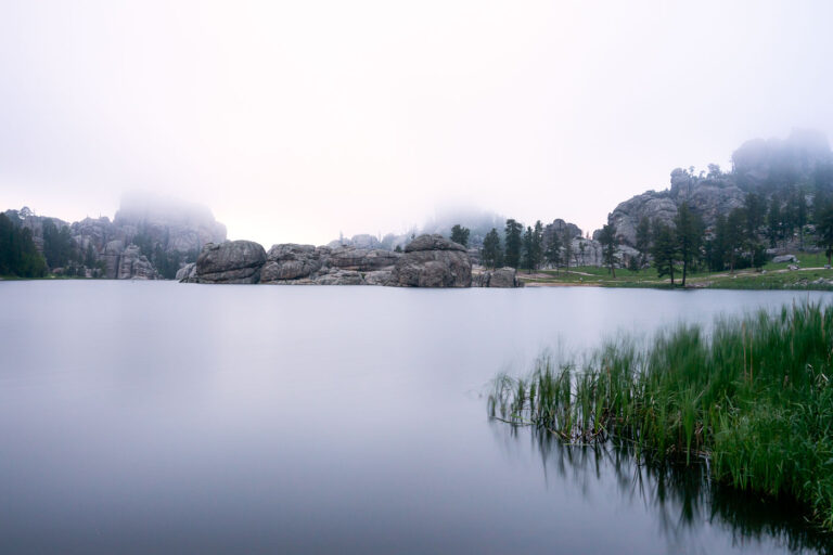 Fog Over Sylvan Lake, Black Hills, South Dakota 2 Early morning fog drifts across Sylvan Lake in the Black Hills of South Dakota, softening the granite spires and reflecting still light off the water’s surface. Created in 1891 by Theodore Reder’s dam, the lake sits at the head of the scenic Needles Highway and is surrounded by granite formations that attract climbers, photographers, and hikers. On this mist-covered morning, visibility narrows to the immediate shoreline, turning the familiar outcrops into shadowed silhouettes. The calm water and diffused light capture a rare quiet moment in one of Custer State Park’s most iconic and photographed landscapes.