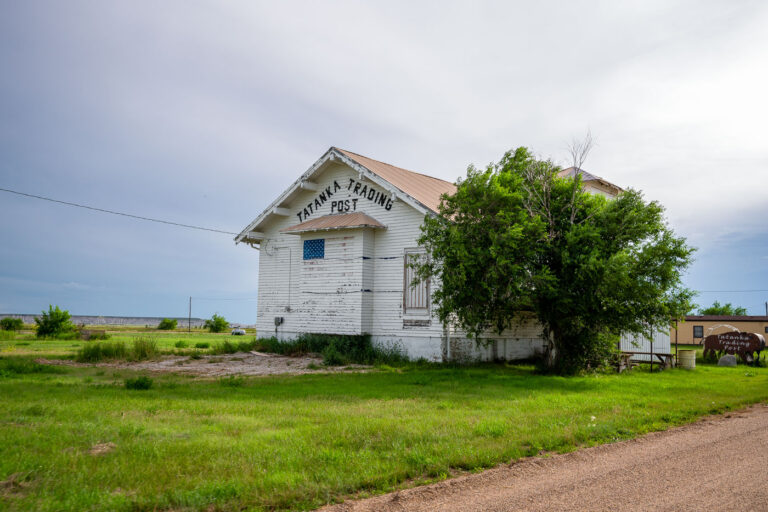Tatanka Trading Post, Scenic, South Dakota 3 The Tatanka Trading Post in Scenic, South Dakota, stands as a weathered relic of roadside commerce along the gateway to Badlands National Park. Once a small but lively hub for travelers and locals, the post’s peeling white paint and simple gabled design speak to decades of wind, sun, and changing times on the Great Plains. The name Tatanka—Lakota for “bison”—reflects the deep cultural ties of the region’s Indigenous heritage and the area’s rugged frontier identity. Today, the building endures as a quiet landmark of rural South Dakota’s layered history.