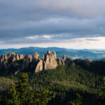 The Needles at Sunset, Black Hills, South Dakota 3 Evening light breaks through heavy clouds to illuminate the Needles, a cluster of towering granite spires rising above dense pine forest in the Black Hills of South Dakota. The sharp pinnacles, part of Custer State Park’s Cathedral Spires formation, are composed of eroded granite that dates back more than a billion years, sculpted by time and weather into their distinctive vertical forms. This region, accessible by the winding Needles Highway, is both a geological marvel and a spiritual landscape—its rugged skyline long revered by the Lakota Sioux and later celebrated by photographers, climbers, and travelers drawn to the surreal beauty of South Dakota’s mountain heartland.