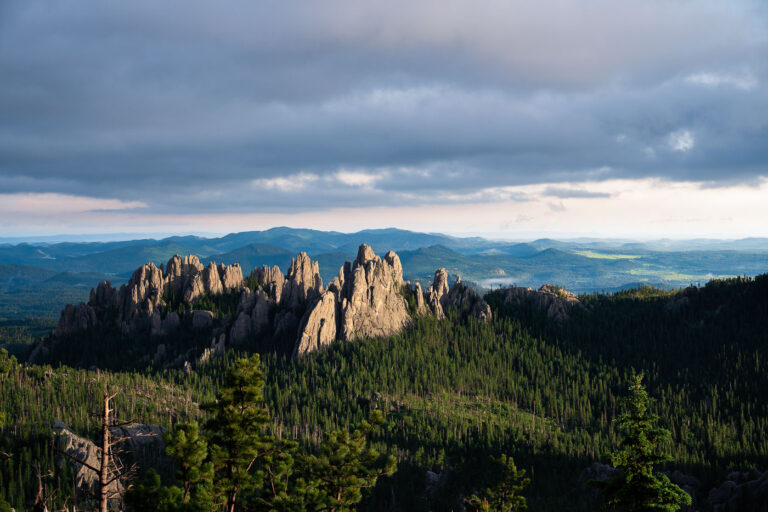 The Needles at Sunset, Black Hills, South Dakota 1 Evening light breaks through heavy clouds to illuminate the Needles, a cluster of towering granite spires rising above dense pine forest in the Black Hills of South Dakota. The sharp pinnacles, part of Custer State Park’s Cathedral Spires formation, are composed of eroded granite that dates back more than a billion years, sculpted by time and weather into their distinctive vertical forms. This region, accessible by the winding Needles Highway, is both a geological marvel and a spiritual landscape—its rugged skyline long revered by the Lakota Sioux and later celebrated by photographers, climbers, and travelers drawn to the surreal beauty of South Dakota’s mountain heartland.