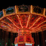 Brightly lit with rows of orange and yellow bulbs, a vintage-style swing carousel glows against the night sky at a fairground. The ornate ride features hand-painted panels depicting alpine landscapes and traditional figures, creating a striking contrast between old-world artistry and modern amusement park energy.