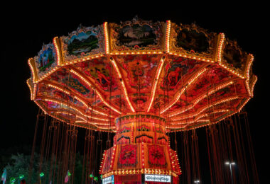 Brightly lit with rows of orange and yellow bulbs, a vintage-style swing carousel glows against the night sky at a fairground. The ornate ride features hand-painted panels depicting alpine landscapes and traditional figures, creating a striking contrast between old-world artistry and modern amusement park energy.