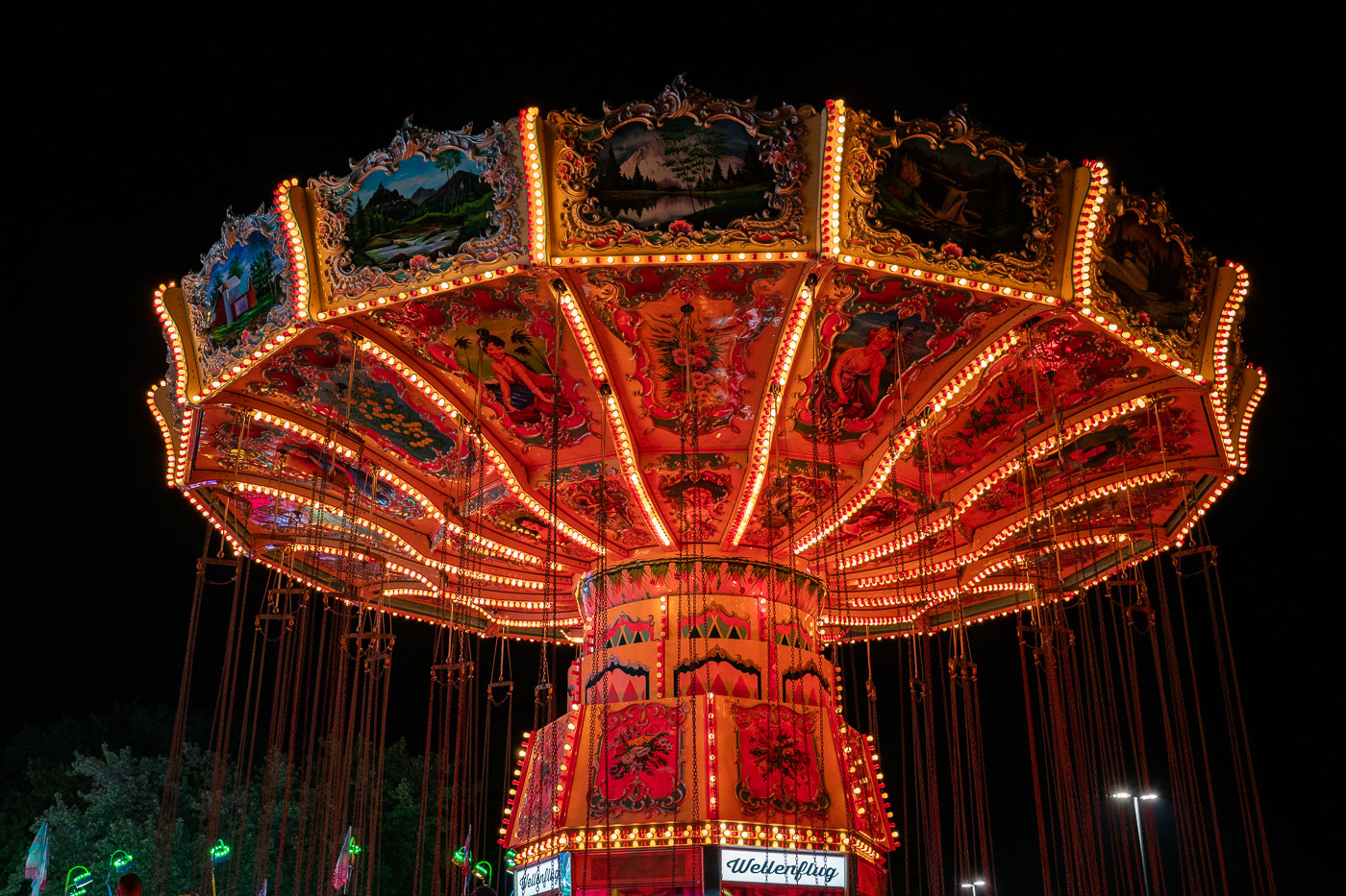 Brightly lit with rows of orange and yellow bulbs, a vintage-style swing carousel glows against the night sky at a fairground. The ornate ride features hand-painted panels depicting alpine landscapes and traditional figures, creating a striking contrast between old-world artistry and modern amusement park energy.