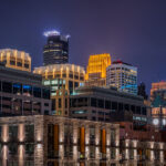 A night view of downtown Minneapolis glows with vibrant hues from its illuminated skyline. The IDS Center and Wells Fargo Tower stand prominently among the city’s modern high-rises, their lights reflecting the urban energy below. New residential buildings in the foreground contrast with the historic clock tower in the distance, highlighting the city’s mix of old and new architecture.